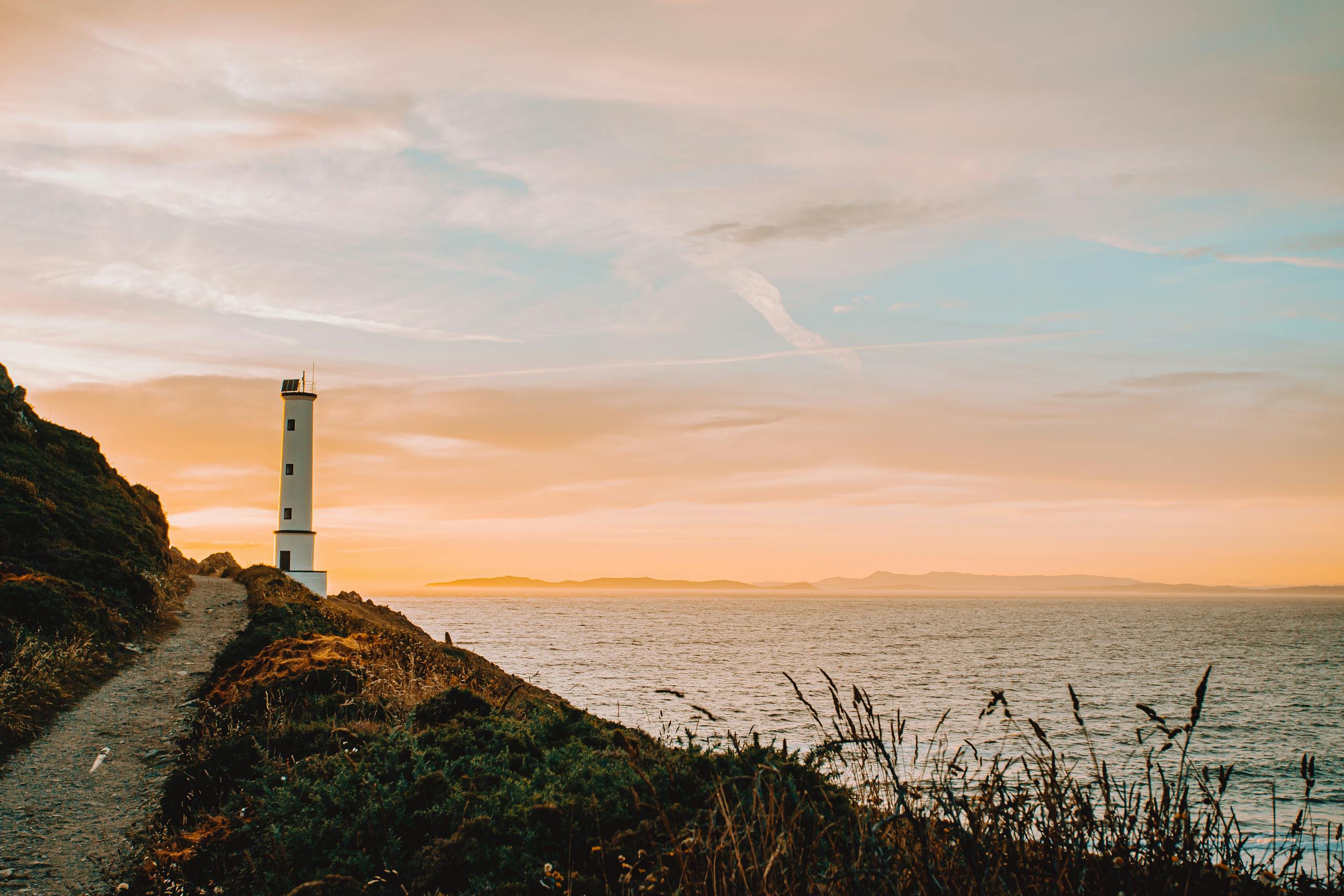 Beacon on mount with narrow path near rippled sea under bright cloudy sky at sunset